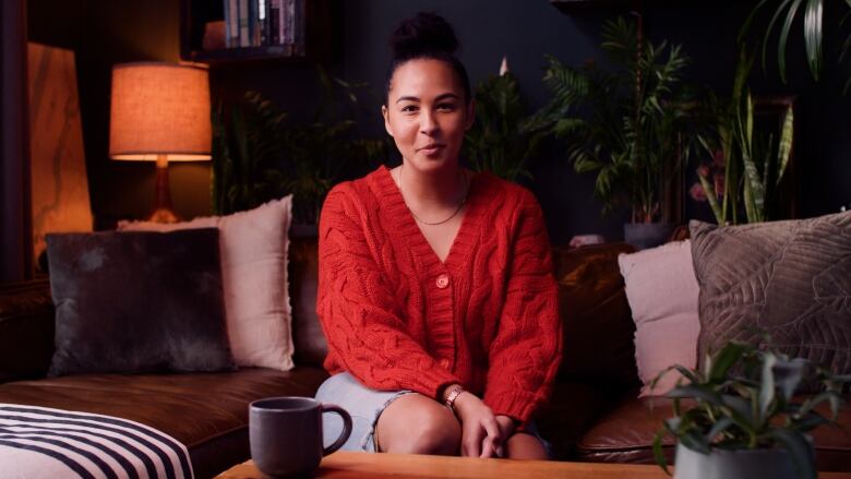 A young woman wearing a red sweater sits on a couch and looks directly at the camera.