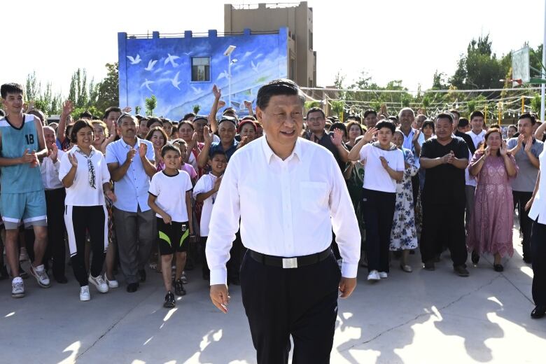 A man in a white shirt and black pants stands in front of a crowd of adults and children.