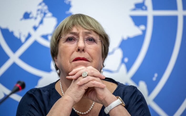 A woman wearing glasses clasps her hands below her chin while sitting at a microphone. 