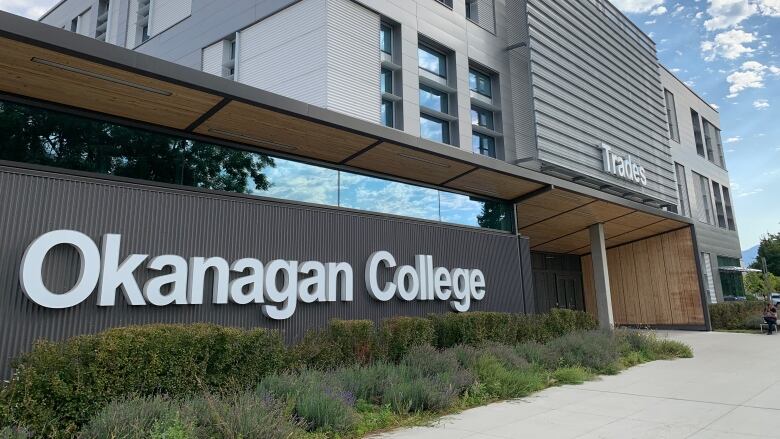 A low-slung, glass-walled building with the sign, 'Okanagan College' connected to a taller grey structure flanked by windows with the world Trades above the entrance.