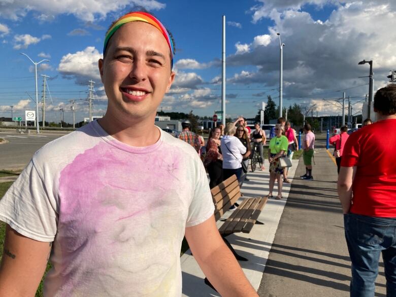 A man poses for a portrait. He's outside in a parking lot with other people behind him. He's wearing a tie-dyed shirt and a rainbow headband.