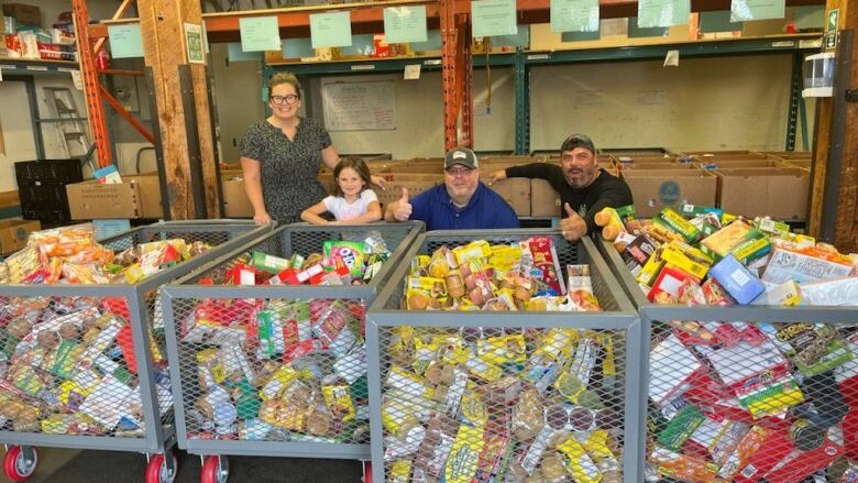 Four people pose behind large, metal bins filled with food in a warehouse.