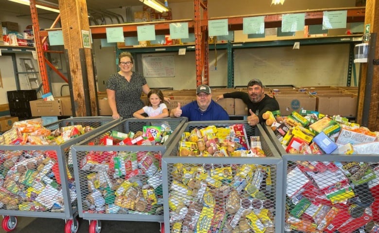 Four people pose behind large, metal bins filled with food in a warehouse.