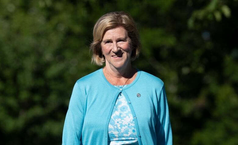 A woman in a blue sweater smiles with trees in the background.