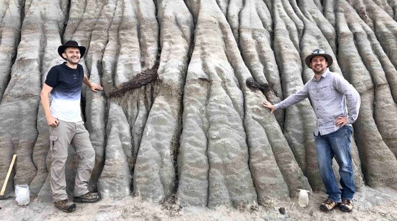 Two smiling men stand beside the fossilized tail of a hadrosaur as it pokes from a cliff face in Dinosaur Provincial Park.