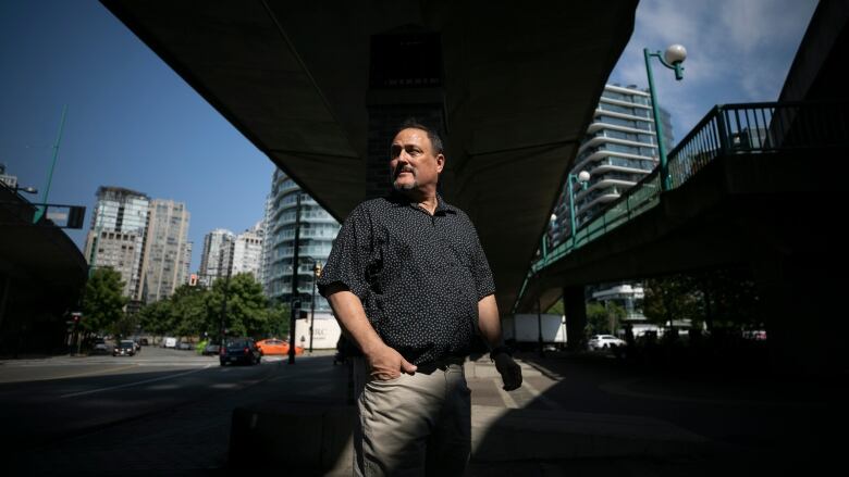 A man in a black patterned shirt stands beneath an overpass in Vancouver.