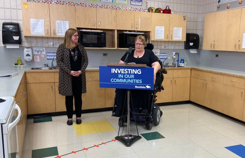 A woman in a wheelchair speaks at a podium in a classroom kitchen.