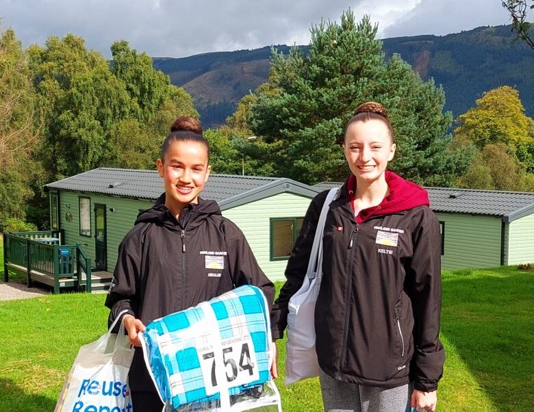 Two girls smile in front of small caravans. One of them is carrying a kilt in a plastic bag. They are both wearing shirts that read 'Highland Dancer' and the B.C. flag.