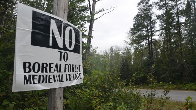 A sign reading 'No to Boreal Forest Medieval Village' is nailed to a tree along a forested road leading to Kenogami Lake. 