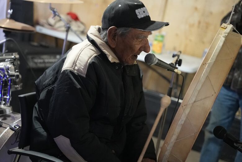 An Innu man sings into a microphone while playing a traditional drum.