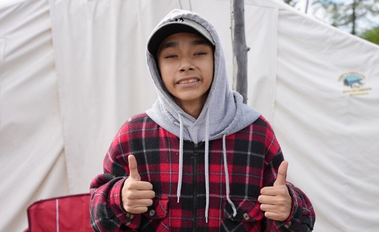 A young Innu dressed in a red plaid hoodie and a ballcap, smiles and gives the double thumbs up in front of an Innu tent.