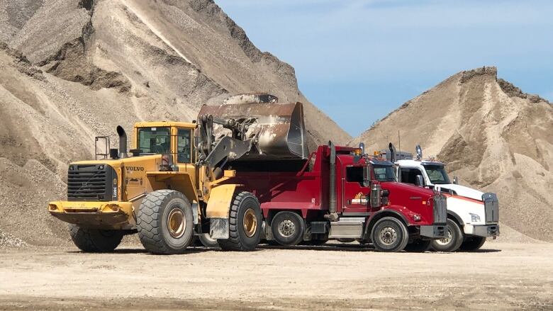 A tractor dumps a load of crushed stone into a truck, with a big mountain of crushed stone in the background.