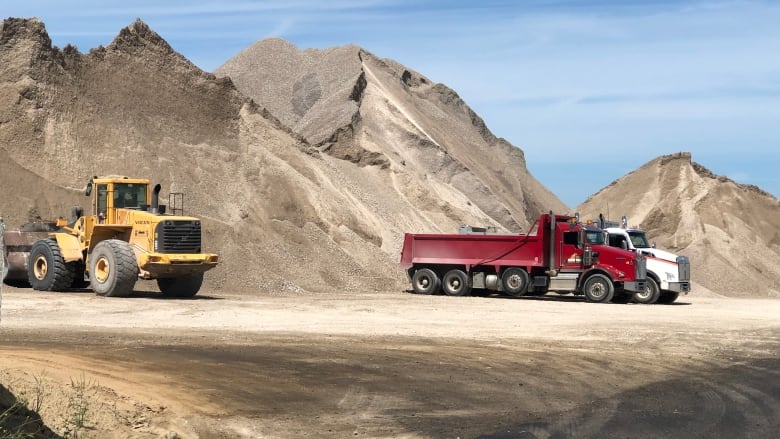 A tractor is seen with two dump trucks against a backdrop of a giant pile of crushed stone.