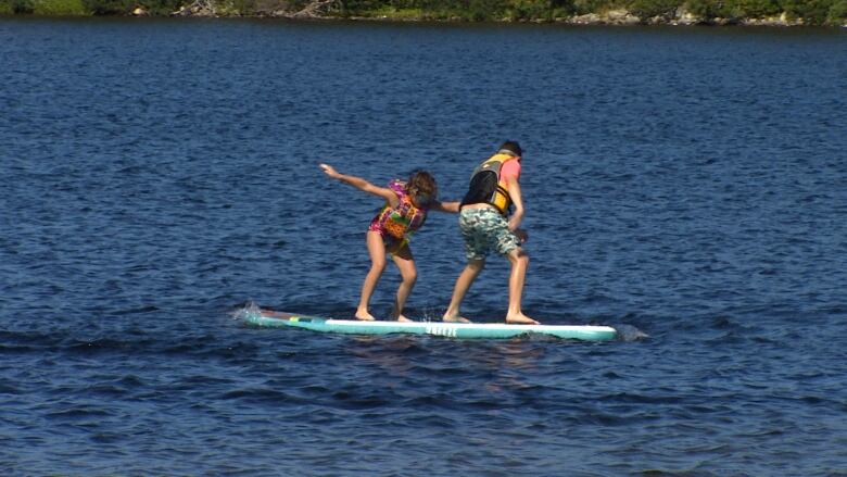 Two children stand on a paddle board in a pond. 