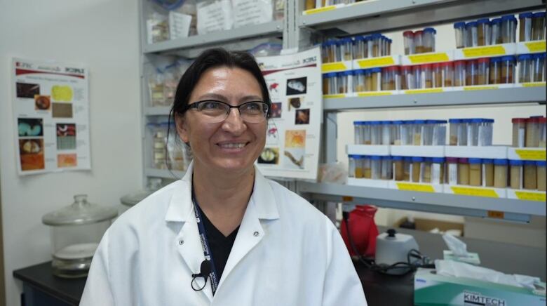 A woman in a white lab coat stands in front of a shelf filled with jars. 