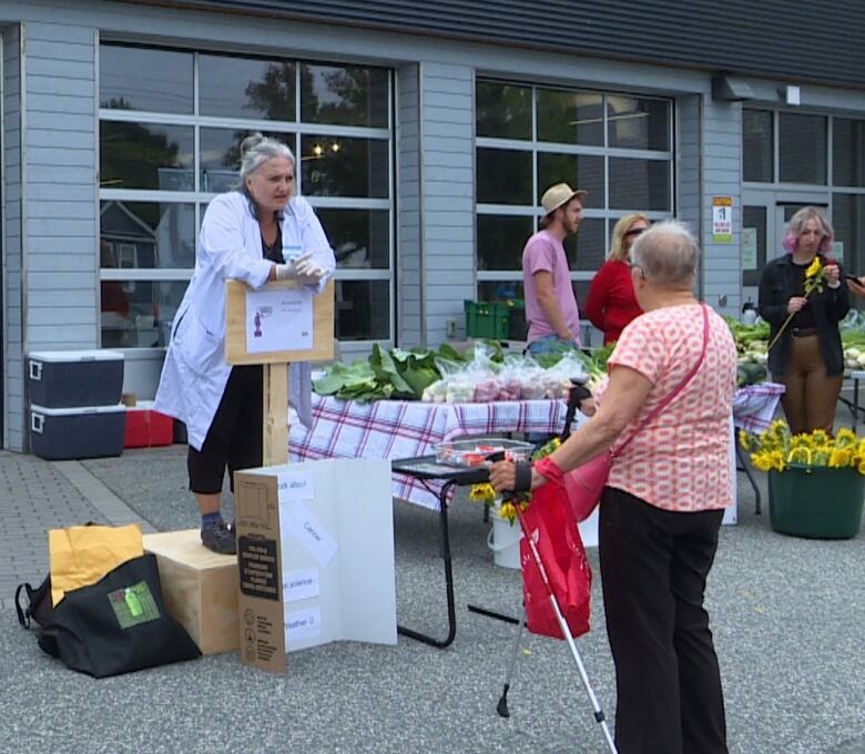 A woman in a lab coat stands on a wooden soapbox, leaning on a sign in front of her that reads Sevtap Savas. She is looking at an older woman in front of her.