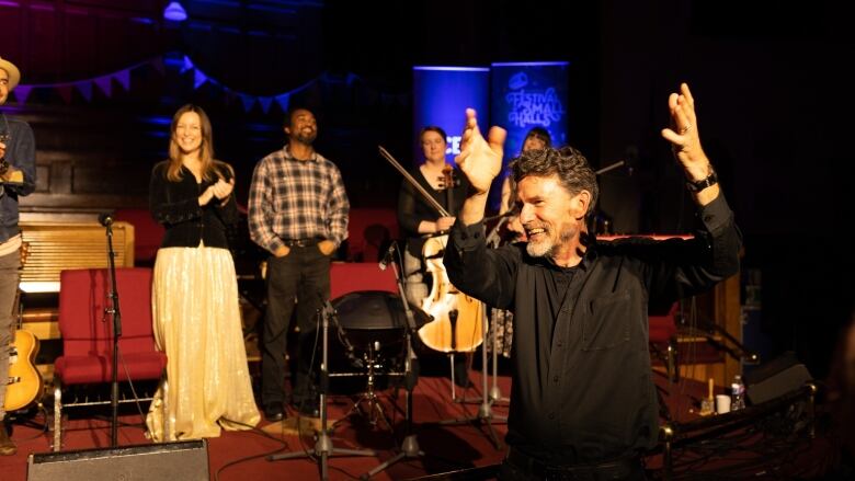 A man is seen with a large smile on his face and arms lifted. He's standing in front of a stage on which a group of musicians stand.