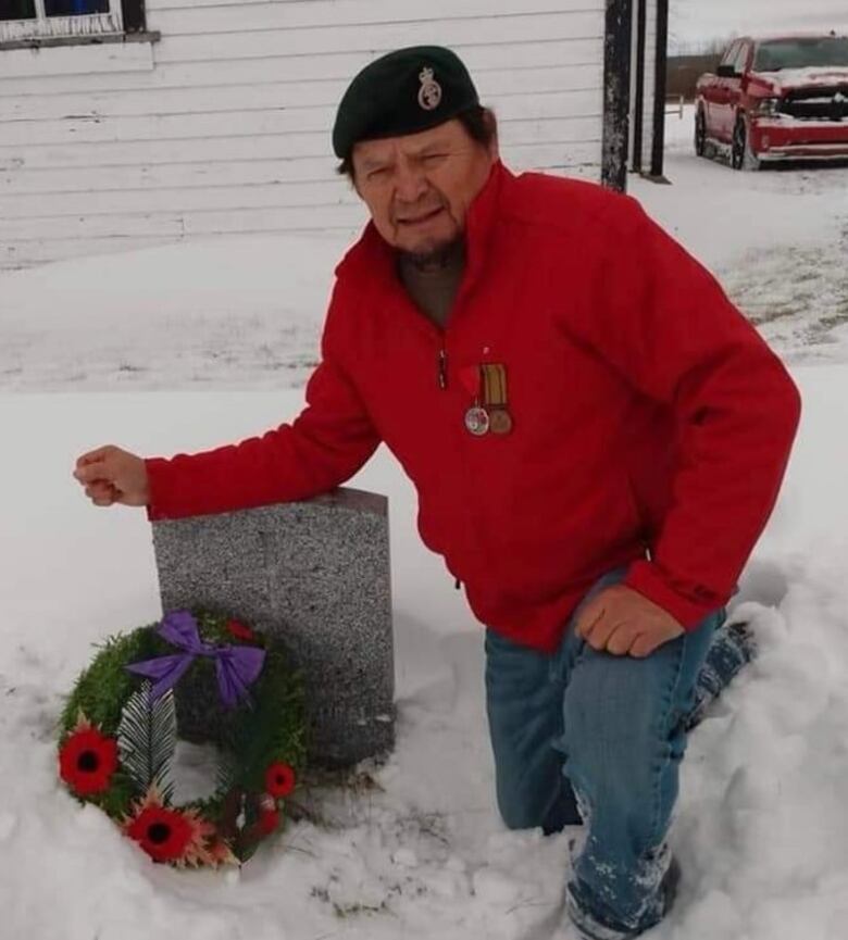 A man in a red jacket kneeling in the snow beside a tombstone with a wreath laid on it. 