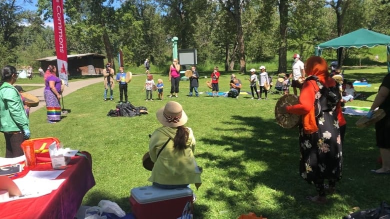 People of all ages stand in a circle in a park on a sunny day holding hand drums.