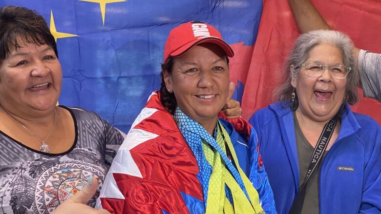 Three smiling people. Person on left is giving thumbs-up, person in centre wears a star blanket and many medals.