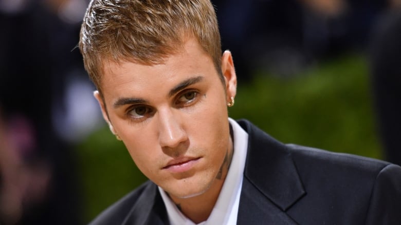 A young man wearing a suit with a tattoo visible on his neck leans in toward the camera. 