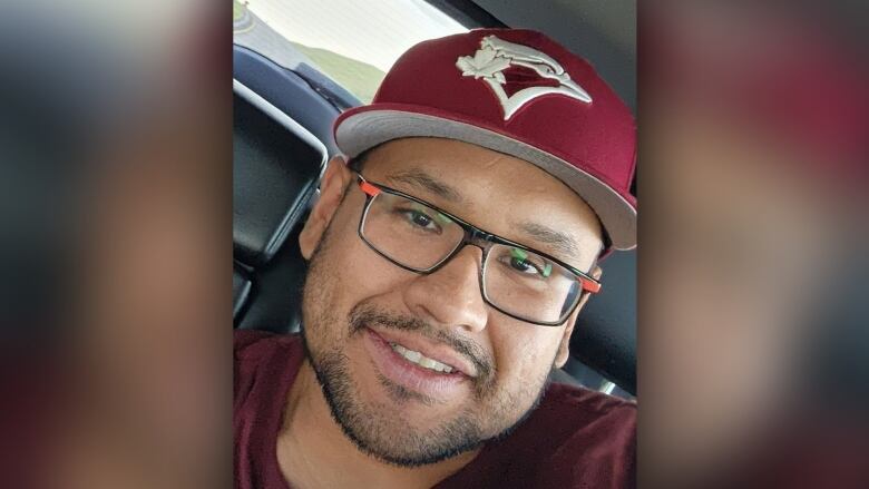 A man with a maroon cap and glasses smiles while sitting in a vehicle.