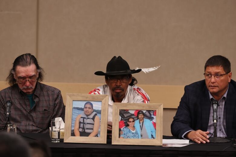 Three men sit at a conference table, on which sit framed photos of three other people. 