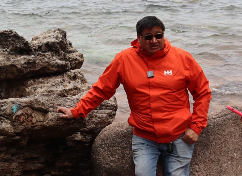 A man in a bright orange rain jacket leans on a rocky outcropping next to the ocean.