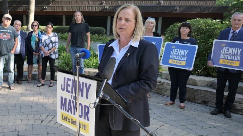 A woman wearing a suit speaks at a podium in front of a line of people, some of them holding signs saying 