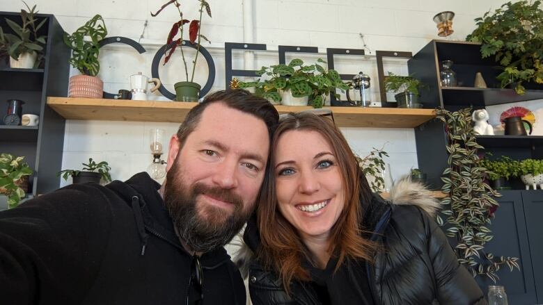 A husband and wife smile in front of a sign that reads 'COFFEE'. The man has black hair and a full beard. The woman has blue eyes and red-streaked hair.