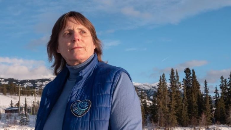 A woman wearing a vest stands in front of a snowy landscape, looking concerned.