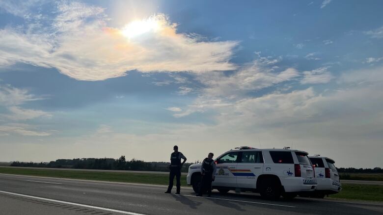 Two RCMP officers stand near two RCMP vehicles on a highway under a partially coudly sky.