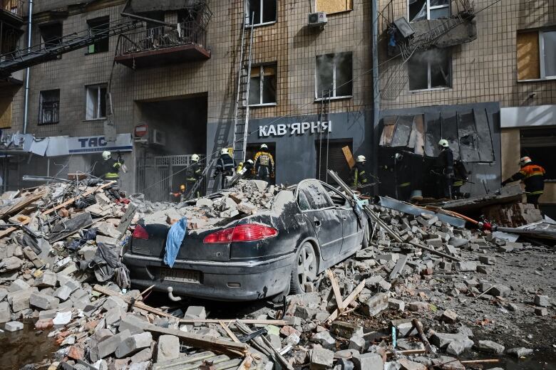 A damaged car, apartment building and rubble are shown on a street.