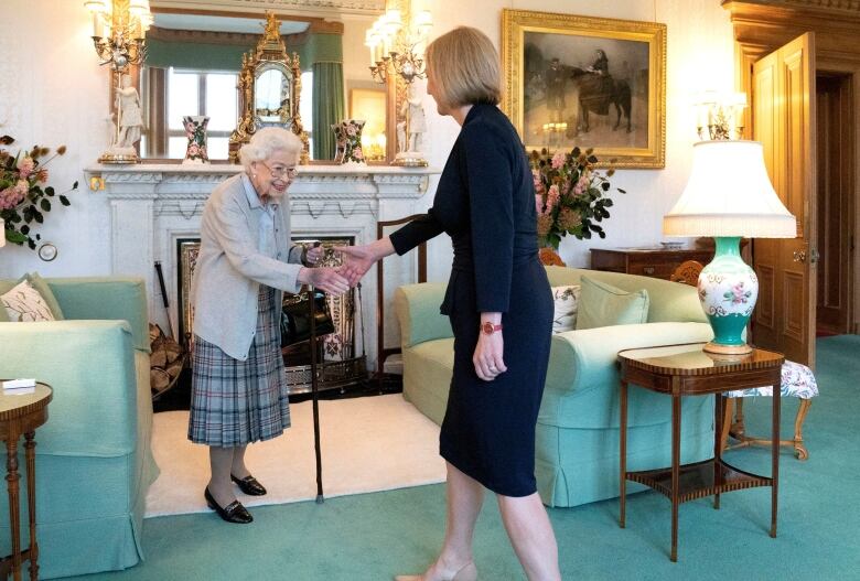 A person carrying a cane shakes hands with another person in a living room.