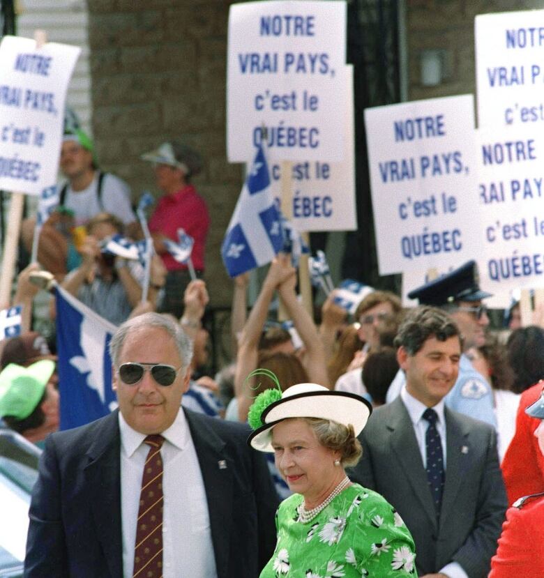 The Queen, in green, in front of protesters holding signs and Quebec flags.