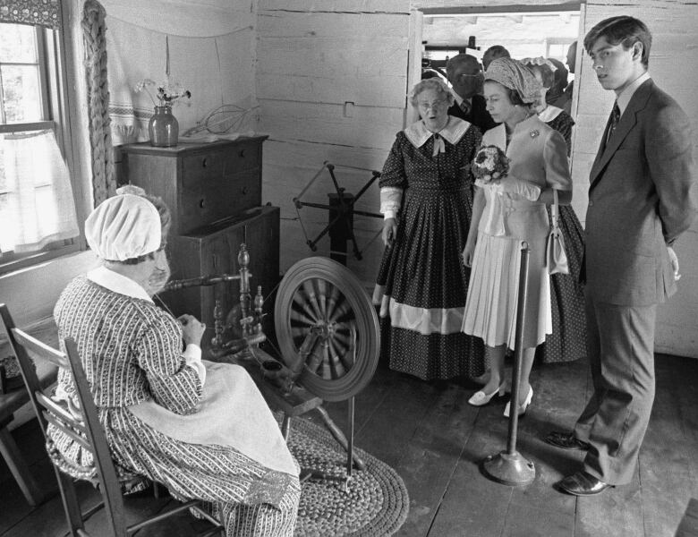 A black-and-white photo of the Queen inside a small wooden building, looking at a spinning wheel.