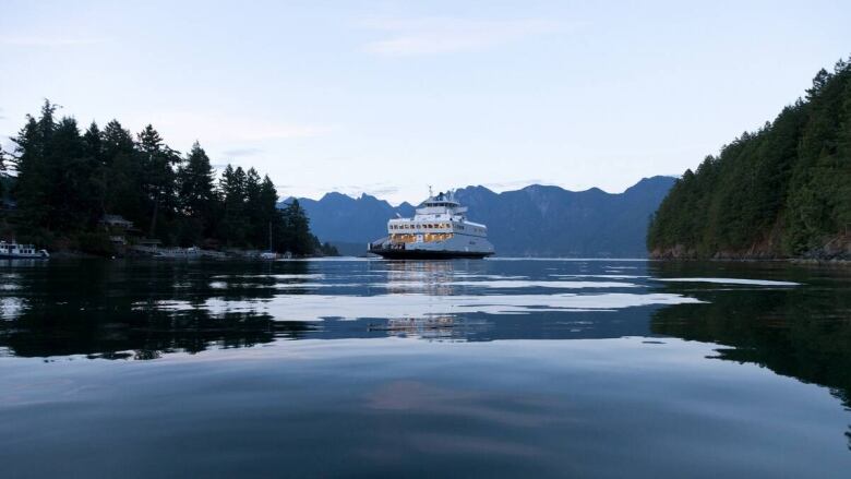 The Bowen Island Ferry on the very calm water of Howe Sound with trees on both sides of the image and mountains in the background. 