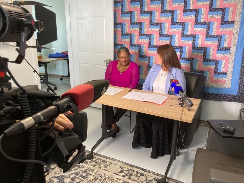 Two women sit at a table with a news camera in the foreground.