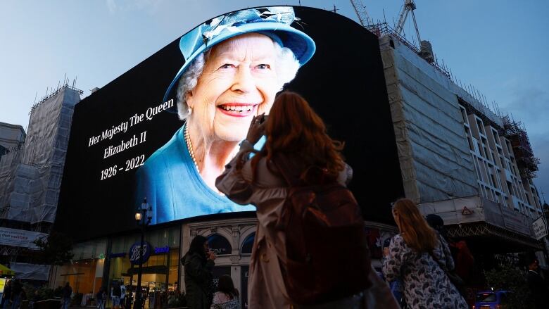 People look up at an image of the Queen on a large pixelboard over a city square. 