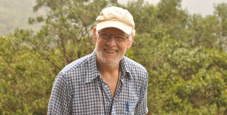 A smiling man in a ball cap stands in front of trees.