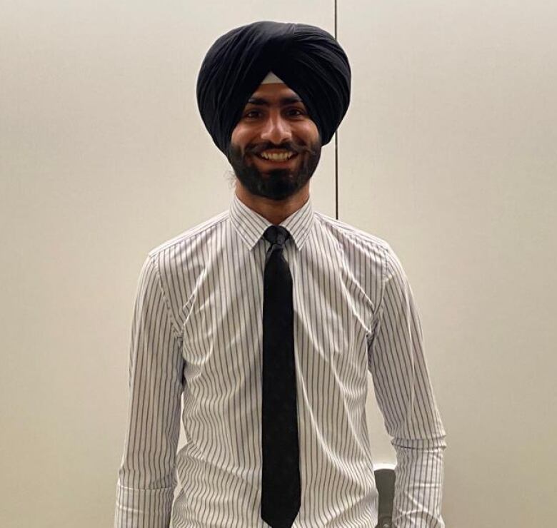 Karan Singh smiles in front of a white wall. He is wearing a long-sleeved shirt with a black tie, and is wearing a black turban.