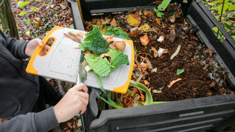 A person's hands are shown holding a cutting board with food scraps in one hand, and a knife in the other, being used to scrape the scraps into a rectangular bin filled with soil and other food scraps.