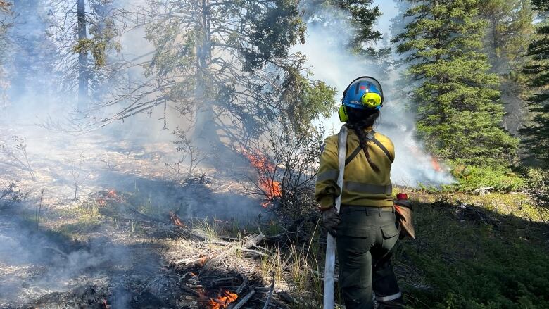 A firefighter is in the woods, holding a hose. The hose is slung over her left shoulder. To her left, there are charred trees and brush with open flames. The burning flames are creating clouds of smoke.