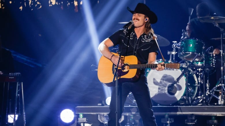 A mean wears a cowboy hat and holds a black guitar while singing on stage with a drum kit behind him.