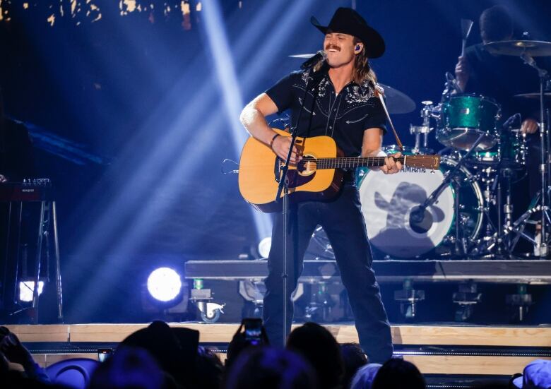 A mean wears a cowboy hat and holds a black guitar while singing on stage with a drum kit behind him.