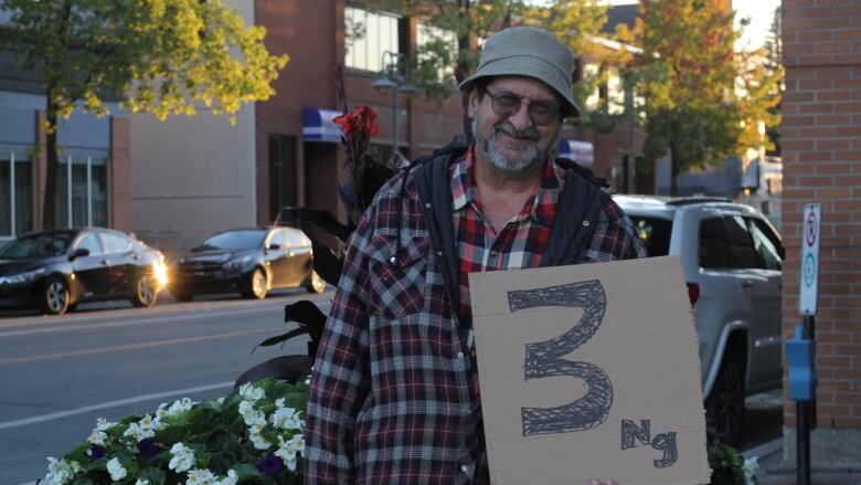 A man in a bucket hat, wearing a plaid jacket, holds a sign that says 3 ng.