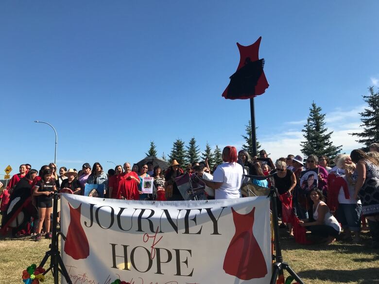 A monument of a red dress and black silhouette is surrounded by friends and family of missing and murdered Indigenous women and girls