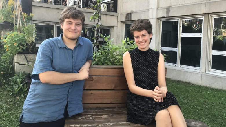 Two students sit outside on a bench in front of a grey building school.