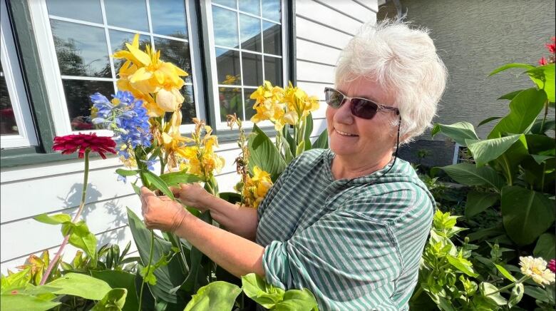 A woman in a green striped shirt with white hair holds a blue flower against a yellow flower.