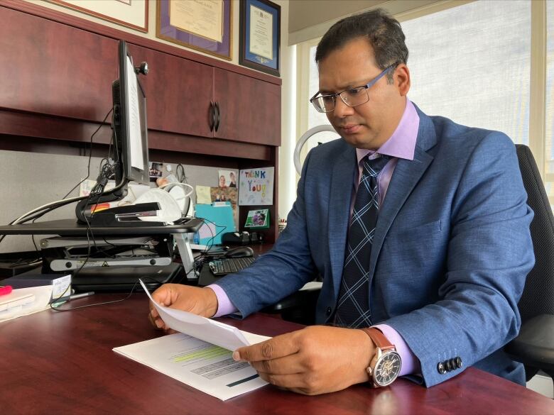 A man in a suit sitting at a desk, looking at some papers he's holding.